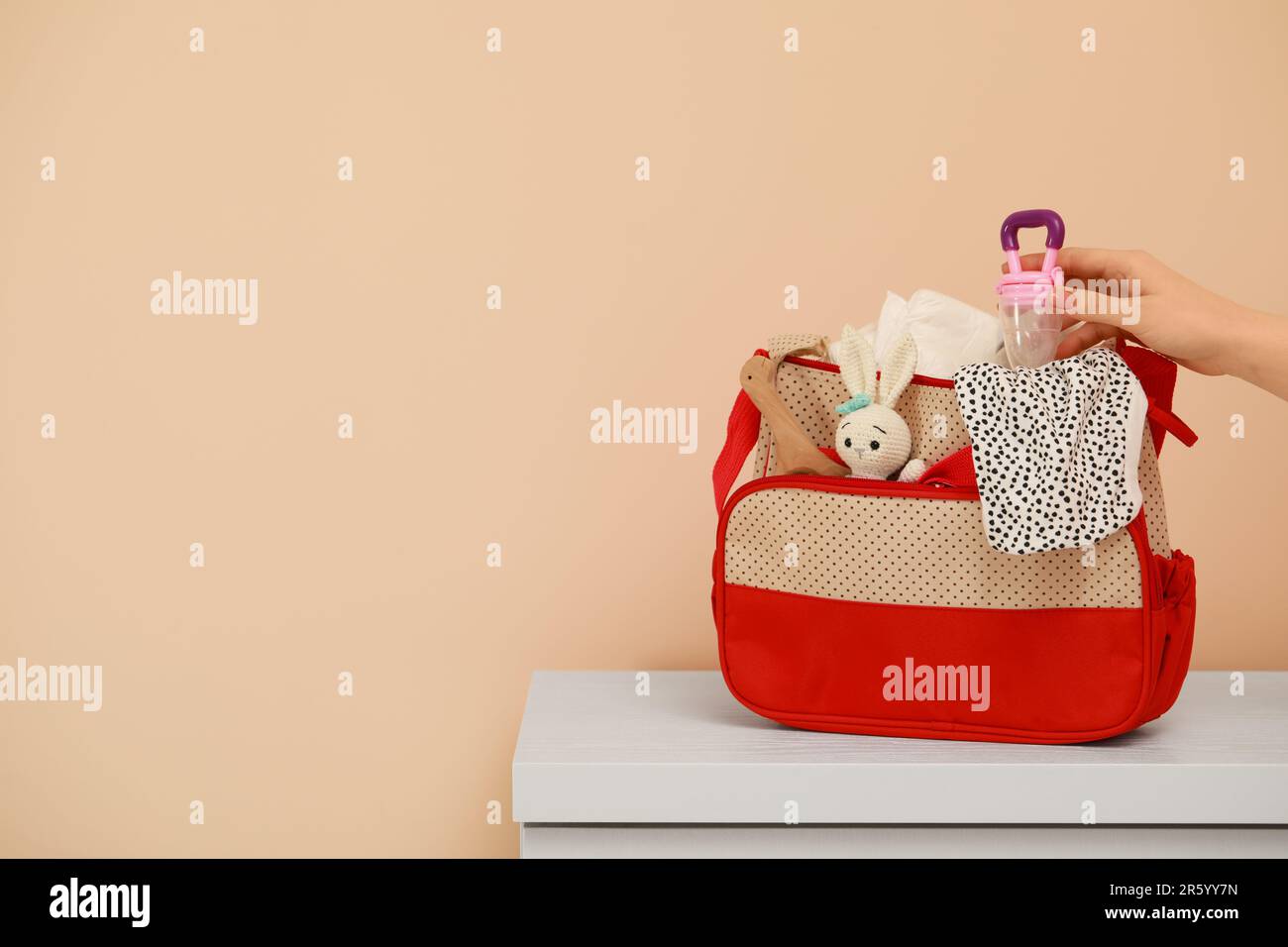 Woman putting empty nibbler into mother`s bag at white table near beige wall, closeup. Space for text Stock Photo